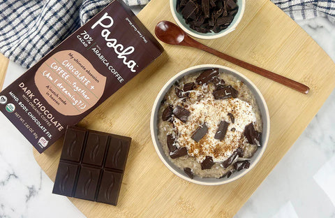 A white bowl of oats and broken pieces of chocolate on top rests on a wooden cutting board sitting on a marble countertop with a 70% bar resting beside the bowl