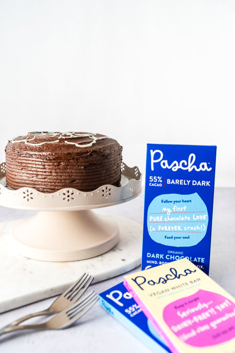 A chocolate cake rested on top of a pink cake stand with some forks on the white counter with a 55% Pascha chocolate bar propped up beside the cake