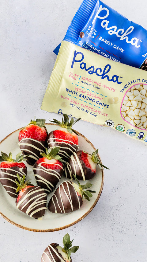 Chocolate covered strawberries sitting on a round white plate resting on a marble surface with Pascha's Organic White and 55% Barely Dark Chocolate Chips on the surface beside.