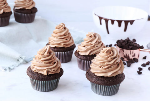 Chocolate cupcakes resting on a marble countertop with a white bowl in the background and melting chocolate dripping down the side. there is also a rose gold measuring cup filled with dark chocolate chips resting on the counter