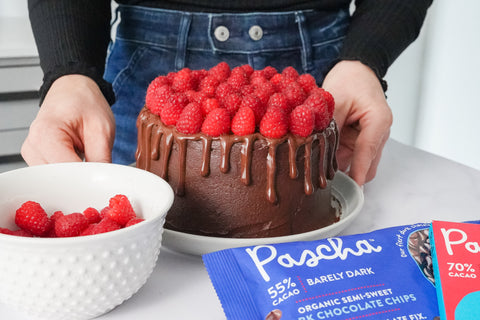 The torso of a person wearing a black long sleeve shirt and blue jeans is placing a chocolate cake topped with raspberries resting on a white plate onto a marble countertop. There is a white bowl of raspberries and in the front of the image is a bag of 55% barely dark chocolate chips bag and a 70% chocolate bar.