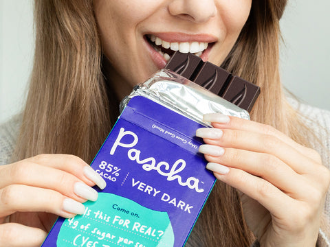 A close up shot of a woman with light brown hair biting a Pascha Chocolate 85% Very Dark Chocolate Bar with the packaging still half on the bare bar