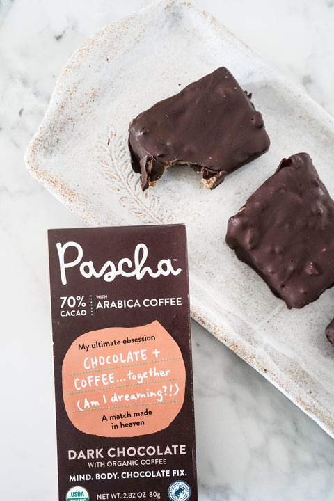 A small chocolate square bar with a bite taken out of it rests beside a full chocolate square bar and both are resting on a plate which sits on a marble surface. There is a 70% Arabica Coffee Chocolate Bar resting on the side of the plate