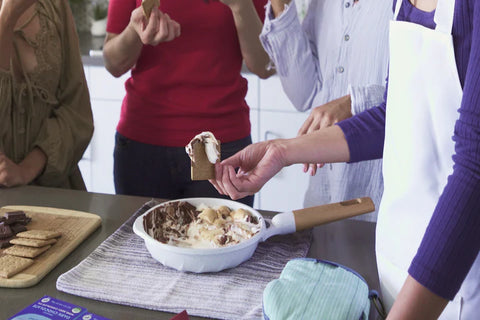 A group of people are standing around a grey countertop dipping graham crackers into a white skillet which contains melted marshmallows and chocolate