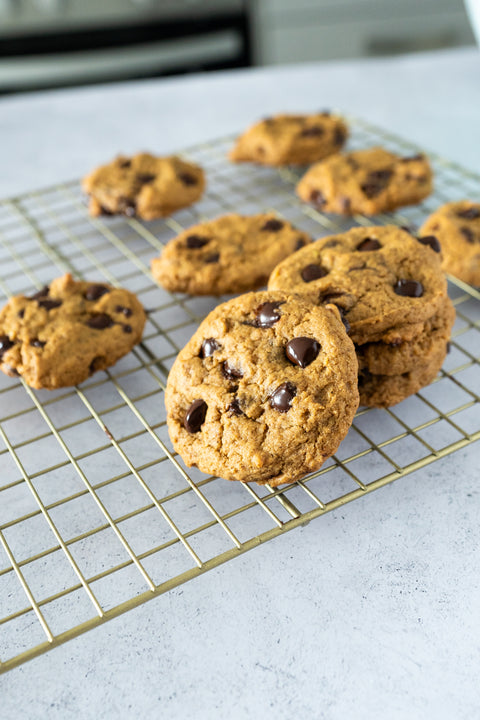 Many chocolate chip cookies are resting on a cooling rack which is sitting on a grey speckled countertop