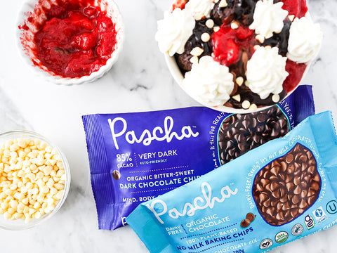 Brownie Ball sundae with vegan whipped cream, strawberry sauce, Pascha mini white chips beside bowls of white chips and strawberry sauce and two bags of Pascha 85% Chips and No Milk Chips 
