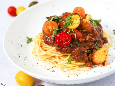 A white bowl filled with angel hair pasta with a vegan chocolate sauce topped with halved cherry tomatoes on top and some parsley to garnish