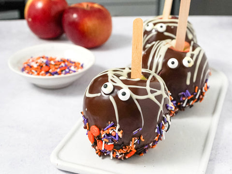 Three chocolate covered apples rest on a plate with white chocolate drizzle and two googely eyes on each. The bottom of the apples have orange and purple sprinkles across them.