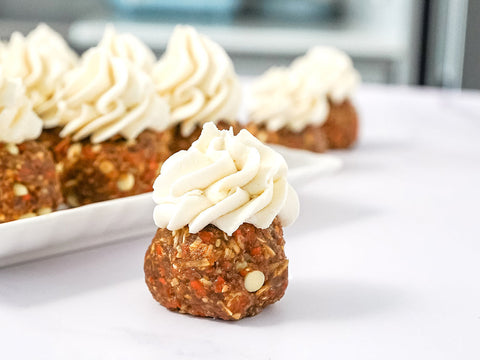 Carrot cake bite topped with swirl of white chocolate cream cheese icing sitting on white counter with plate full of bites in the background.