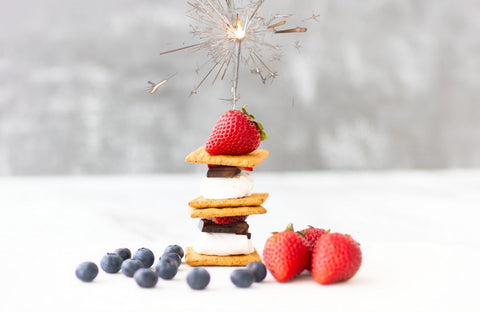 Stack of s'mores sitting on a white surface with blueberries and strawberries around them. There is a single strawberry placed on top of the stack with a lit sparkler in the strawberry.