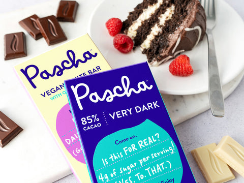 Two Pascha chocolate bars, 85% and Vegan white bar, laying on a marble countertop with a slice of cake on a plate beside the bars