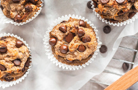 Banana chocolate chip muffins in white cupcake holders placed on parchment paper with a few chocolate chips surrounding the muffins