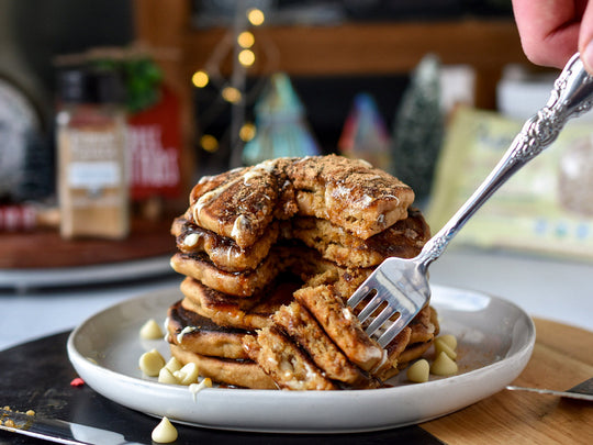 Stack of pancakes on a white plate with a fork sticking in a bite. Pancakes are topped with melted Pascha White Chocolate and sprinkled with White Chocolate Chips.