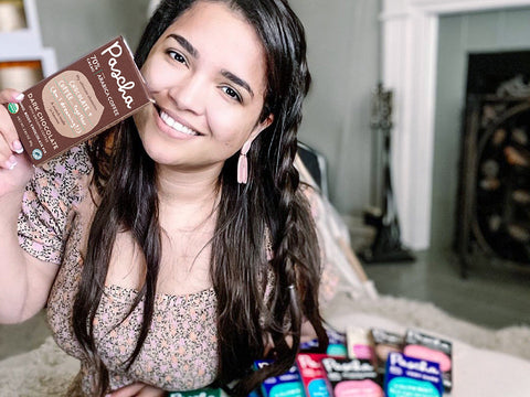 smiling dark haired lady in a brown floral dress holding a Pascha coffee bar sitting on a couch in a living room with a pile of Pascha bars beside her