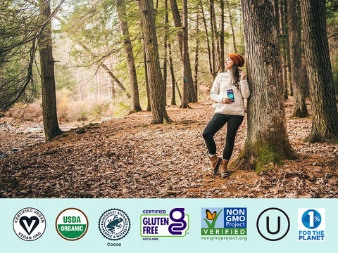 Woman standing in a forest leaning against a tree wearing a red beanie, white sweater, black leggings and brown boots. There is a banner at the bottom of the image with all of Pascha's certifications listed.