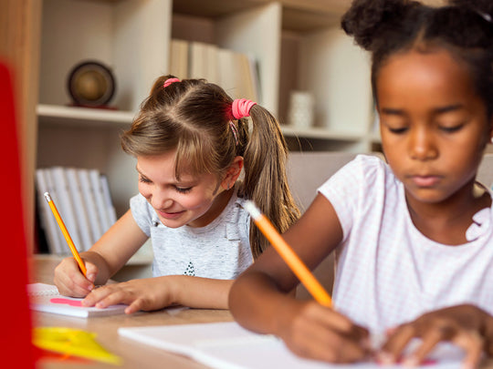 Two young girls happily writing in notebooks with pencils.
