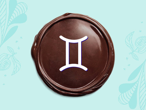 White Gemini sign on a disc of melted dark chocolate on a light teal background.
