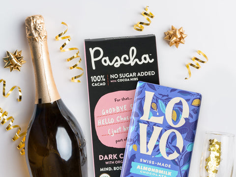 A champagne bottle resting on a white surface beside a Pascha 100% No Sugar Added bar and a LOVO Almondmilk Chocolate bar