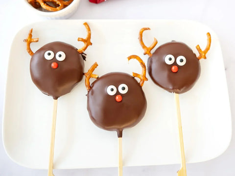 3 Reindeer pops made out of chocolate dipped cookie dough, pretzels and candy eyes on a white plate.