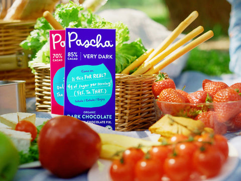 Two Pascha bars leaning against wicker picnic basket surrounded by fresh lettuce, strawberries and tomatoes.