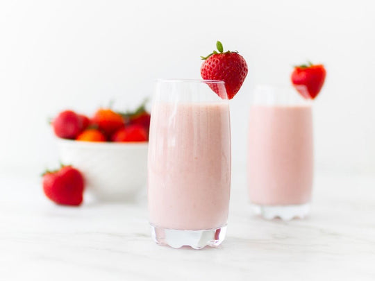 Two glasses of white chocolate strawberry smoothies in tall clear glasses, topped with a whole strawberry and bowl of fresh strawberries in the background.