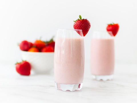 Two glasses of white chocolate strawberry smoothies in tall clear glasses, topped with a whole strawberry and bowl of fresh strawberries in the background.