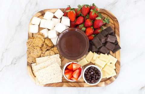 Wooden cutting board with melted chocolate in a bowl in the middle surrounded by snacks such as strawberries, marshmallows, crackers and chocolate bar pieces