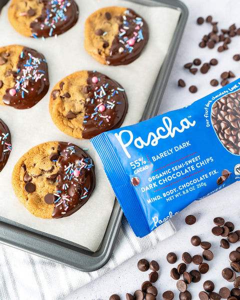 Resting on a grey speckled surface there is a cooking tray with parchment paper lining it with loose chocolate chips surrounding along with a 55% Pascha Chocolate Chip bag. On the parchment paper there are 6 chocolate chip cookies, half dipped in chocolate and sprinkles