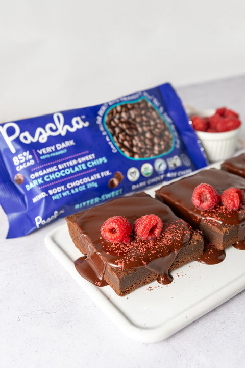 There are three brownies on a white rectangular plate, covered in melted chocolate and raspberries sitting on a white plate with a pack of 85% Chips in the background