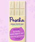The front of Pascha Chocolate's Vegan White Bar with half of the exposed bar sticking out of the top of the packaging on a light pink background with subtle snowflakes.