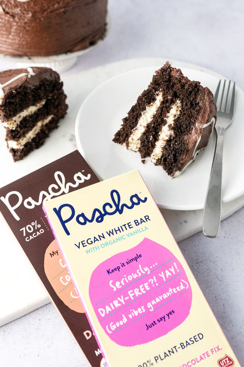 Pascha's Vegan White and 70% Cacao Coffee Bars are laying stacked on top of one another on a grey speckled surface beside a white plate with a fork and slice of chocolate and white filled cake. There is a glimpse of the full cake in the top left corner.