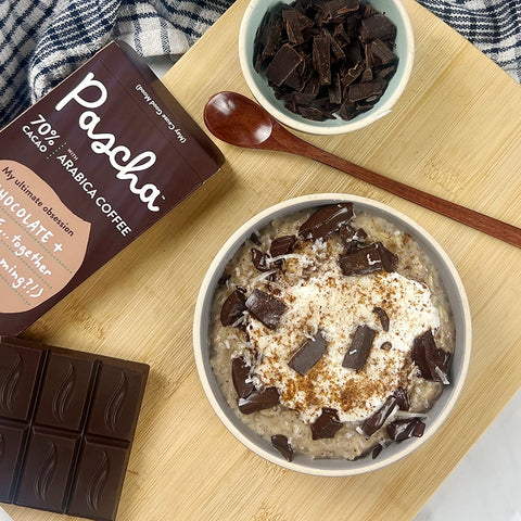 Chocolate banana oat bowl is in a white bowl resting on a wooden board. There is also a bowl of chopped chocolate bar pieces on the board along with a wooden spoon and a bar of 70% Pascha chocolate arabica coffee