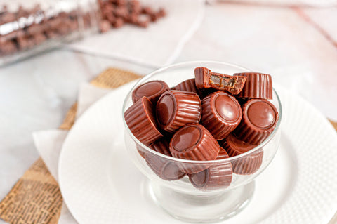 Chocolate covered caramels sitting in a glass bowl on top of a white plate placed on a countertop with a jar spilling out chocolate covered caramels in the background