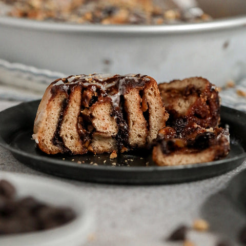 Chocolate hazelnut cinnamon roll cut in half on a black small plate with the tin of the rest of the cinnamon rolls in the backgorund. 