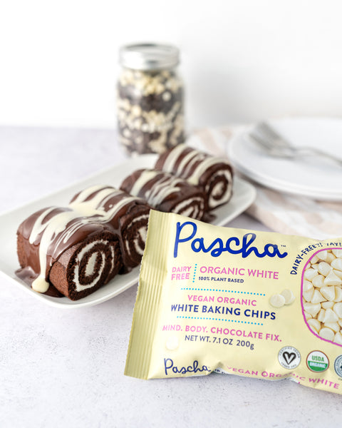 White and Dark chocolate cake rolls are resting on a white rectangular plate with melted white and dark chocolate drizzled on top. There is a White organic vegan baking chips bag resting in front and a clear jar with white and dark chocolate chips is resting in the background 