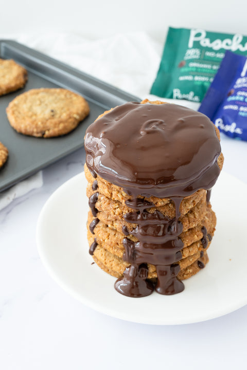 A stack of six chocolate chip cookies rests on a white plate placed on a white table with melted chocolate covering the top cookie and melting down the sides of the other cookies. There are freshly baked cookies resting on a baking sheet in the back along with some Pascha chip bags. 