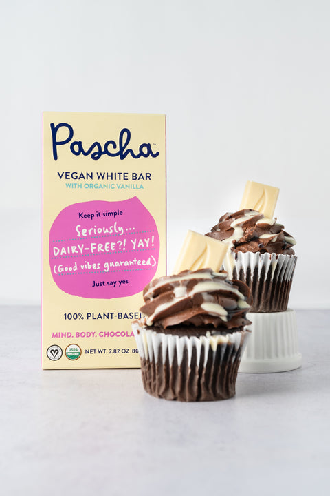 Pascha's Vegan White Bar is standing upright beside two chocolate cupcakes, one of which is on a small white upside down bowl all on a marble surface. Both of the cupcakes have a piece of the white bar pieces in them.