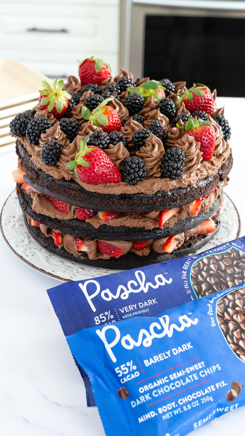 Three layers of chocolate cake with a chocolate icing and fresh fruit on top of each layer form one three layered cake. The cake rests on a white plate sitting on a marble countertop with two dark chocolate bags resting beside teh cake 