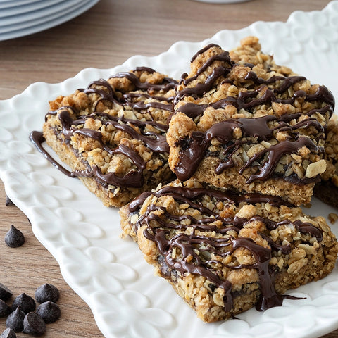 Date and oatmeal squares stacked on a white plate that has white flowers outlining the edge. The plate is on a brown table with a few chocolate chips as well 