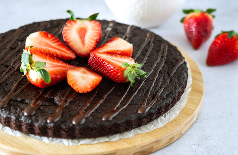 Thin round chocolate cake topped with halved strawberries and a drizzle of melted chocolate sitting on a round wooden platter resting on a grey speckled surface