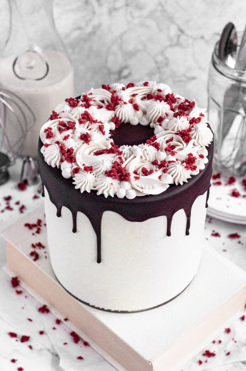 A white cake rests on top of a white book. The cake has drizzled chocolates coming down the sides and is topped with a ring of white icing and some red velvet cake crumbs sprinkled over the cake. Some of the red velvet crumbs are on the book and counter surrounding. There isa. glass of milk in the background along with cutlery in a glass jar. 