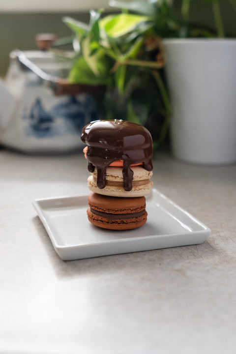 Three macaroons chocolate, caramel and strawberry macaroons are stacked on top of one another, resting on a white square plate and have melted chocolate covering the top strawberry one and drizzle down the sides. There is a plant in the background sitting in a white vase.