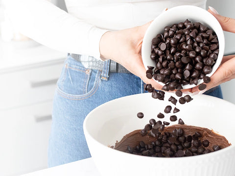 Woman holding a white bowl full of Pascha Chocolate chips and pouring them into another white bowl of more melted chocolate on a white counter.