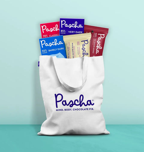 A Pascha Chocolate tote bag which reads Pascha mind. body. chocolate fix. is leaning agains a teal wall with several Pascha products poking out of the top 