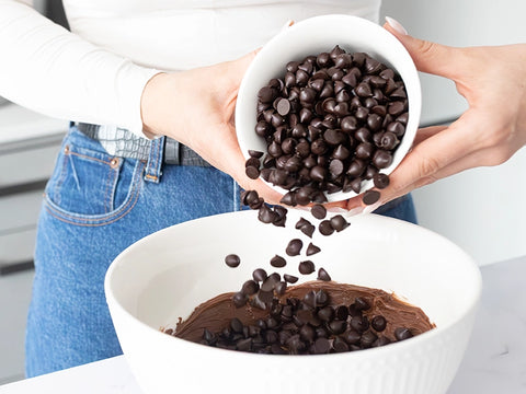 A woman wearing a white long sleeve shirt and blue jeans is pouring a white bowl full of chocolate chips into another larger white bowl of melted chocolate.