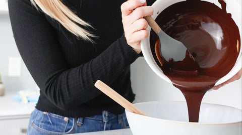 A person wearing a long sleeve black top and denim jeans is pouring a white bowl of melted dark chocolate into another bowl that is resting on a countertop