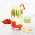 a tall clear glass is resting on a wooden surface with muddled strawberries in the bottom with a layer of milk in the middle and then a layer of matcha on top. There is a thin strawberry slice on the rim of the glass and there are white chocolate chips and full strawberries placed beside the glass. 