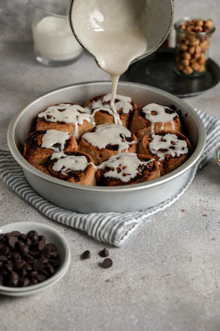 There are eight vegan cinnamon hazelnut rolls in a circular baking dish with a white bowl pouring white icing all over the top of them. They are resting on a grey stripped cloth which is on a grey speckled surface with a glass of milk and clear jar of hazelnuts in the background