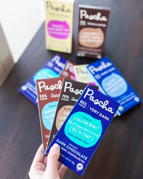 Three Pascha Chocolate bars fanned out in a hand with many more Pascha Chocolate bars placed on a brown table below