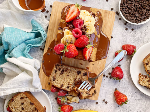 chocolate banana bread topped with fresh strawberries, banana slices, raspberries and chocolate chips rests with melted chocolate poured over top on a wood cutting board which rests on a grey marble countertop surrounded by a white bowl of chocolate chips and a variety of cloths. 